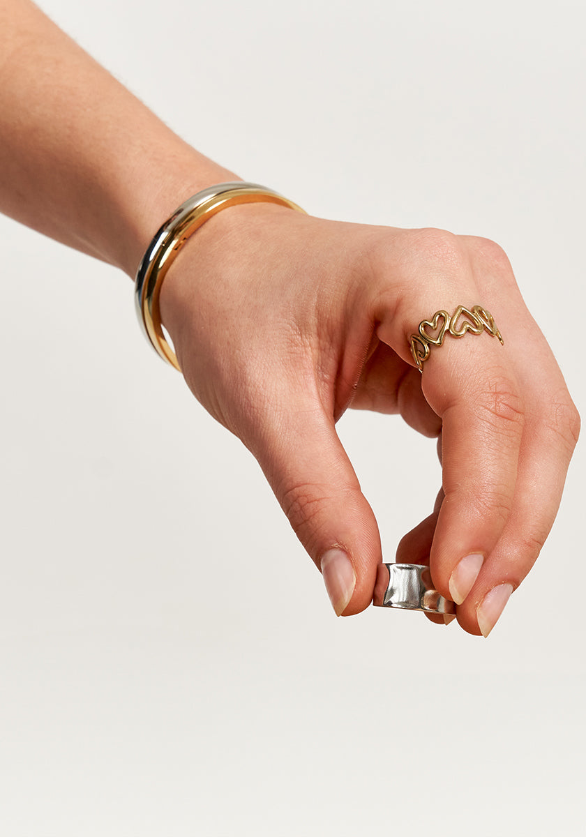 A hand adorned with a gold bracelet and the Princess Goes Hollywood Silberner Ring mit Struktur delicately holds a small, shiny, rectangular object aus Edelstahl 304. The background is plain and light-colored.