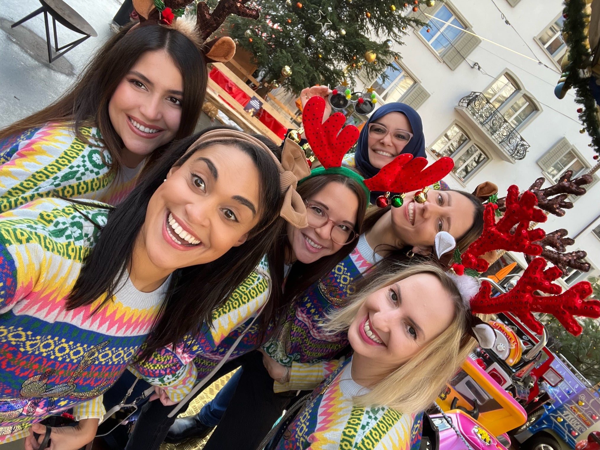 Six women wearing colorful festive sweaters and reindeer antler headbands smile together for a selfie outdoors. Behind them is a decorated Christmas tree and a small colorful train ride, suggesting a holiday celebration in a city square.