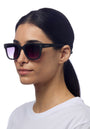 A woman with straight dark hair in a low ponytail wears Okkia square-frame black sunglasses with gradient lenses. She has a neutral expression, is dressed in a plain white shirt, and stands against a solid white background.