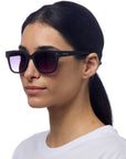 A woman with straight dark hair in a low ponytail wears Okkia square-frame black sunglasses with gradient lenses. She has a neutral expression, is dressed in a plain white shirt, and stands against a solid white background.