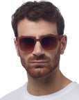 A man with short, wavy brown hair and a trimmed reddish-brown beard wears Okkia unisex dark brown square-frame sunglasses with UV 400-Schutz and a white crew-neck shirt, facing forward against a plain white background.
