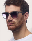 Ein Mann mit kurzen braunen Haaren und gestutztem Bart trägt eine große, dunkle Okkia-Sonnenbrille mit UV-400-Schutz und Square-Frame-Design. Er blickt leicht nach rechts vor einem schlichten, hellen Hintergrund, sein Ausdruck ist neutral.