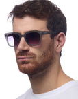 A man with short brown hair and a trimmed beard wears black-framed Okkia sunglasses featuring a square-frame design and a white T-shirt. He faces slightly left against a plain white background with a neutral expression.