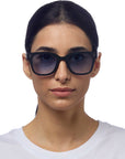 A woman with straight dark hair pulled back wears Okkia oversized black sunglasses with a square-frame design and UV 400 protection. She faces the camera in a white crew-neck shirt, her expression neutral against a plain white background.