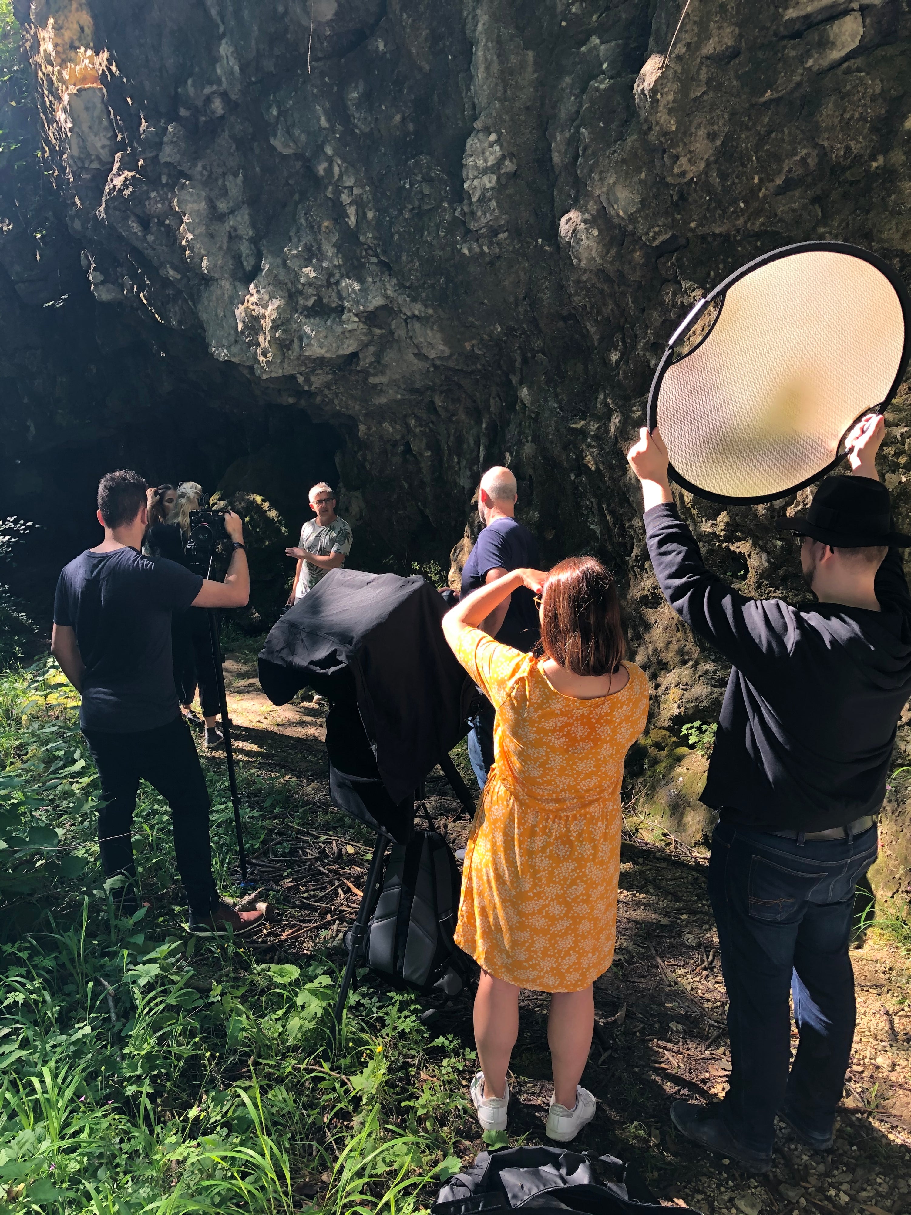 A group of five people conducts an outdoor photoshoot near a rocky cave. One person holds a large reflector, another adjusts a camera, and a woman in a yellow dress stands in front, while a man poses near the cave entrance. Sunlight filters through the trees.
