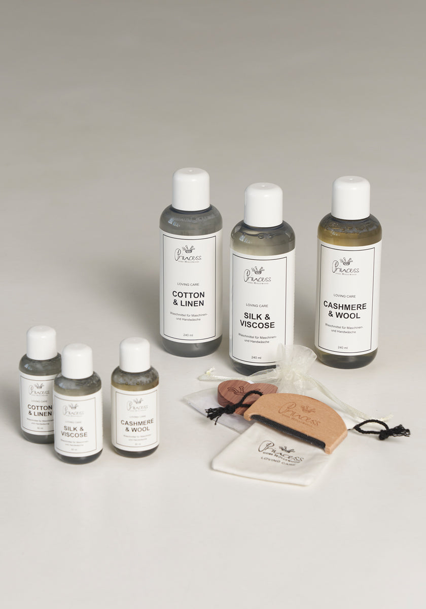 Seven bottles of Princess Goes Hollywood Kaschmir & Wolle Pflegewaschmittel are displayed on a light surface. Labels read Cotton & Linen, Silk & Viscose, and Cashmere & Wool. Nearby are fabric tags, a pouch, and a small brush.