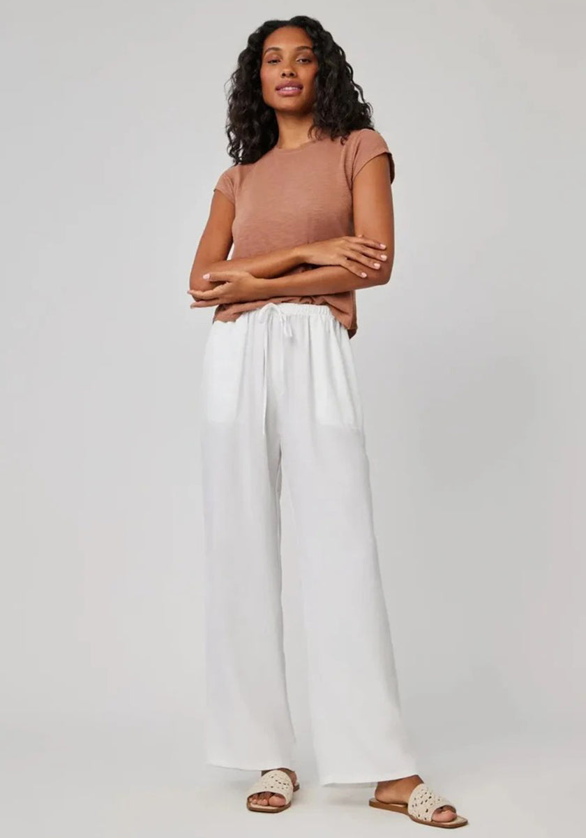 A woman with curly hair stands against a plain background, wearing a Bella Dahl short-sleeve brown top and white pants with an elastic waistband. Beige slide sandals complete her look as she calmly poses with arms lightly crossed.
