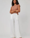 A woman with curly hair stands against a plain background, wearing a Bella Dahl short-sleeve brown top and white pants with an elastic waistband. Beige slide sandals complete her look as she calmly poses with arms lightly crossed.