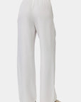 A person is shown from the waist down, wearing Bella Dahls loose-fitting, wide-leg white Lyocell Damenhose with elastischem Bund. The ankle-length pants are paired with open-toed sandals against a plain, light background.