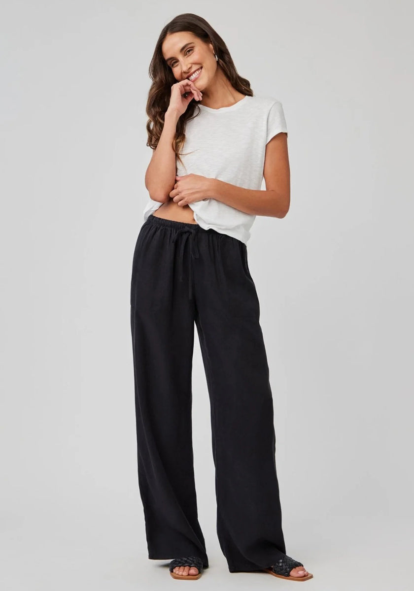 A woman with loose brown wavy hair stands against a light gray background, smiling with one hand on her chin. She wears a Bella Dahl white short-sleeved T-shirt, white Lyocell pants with an elastic waistband, and black slide sandals.