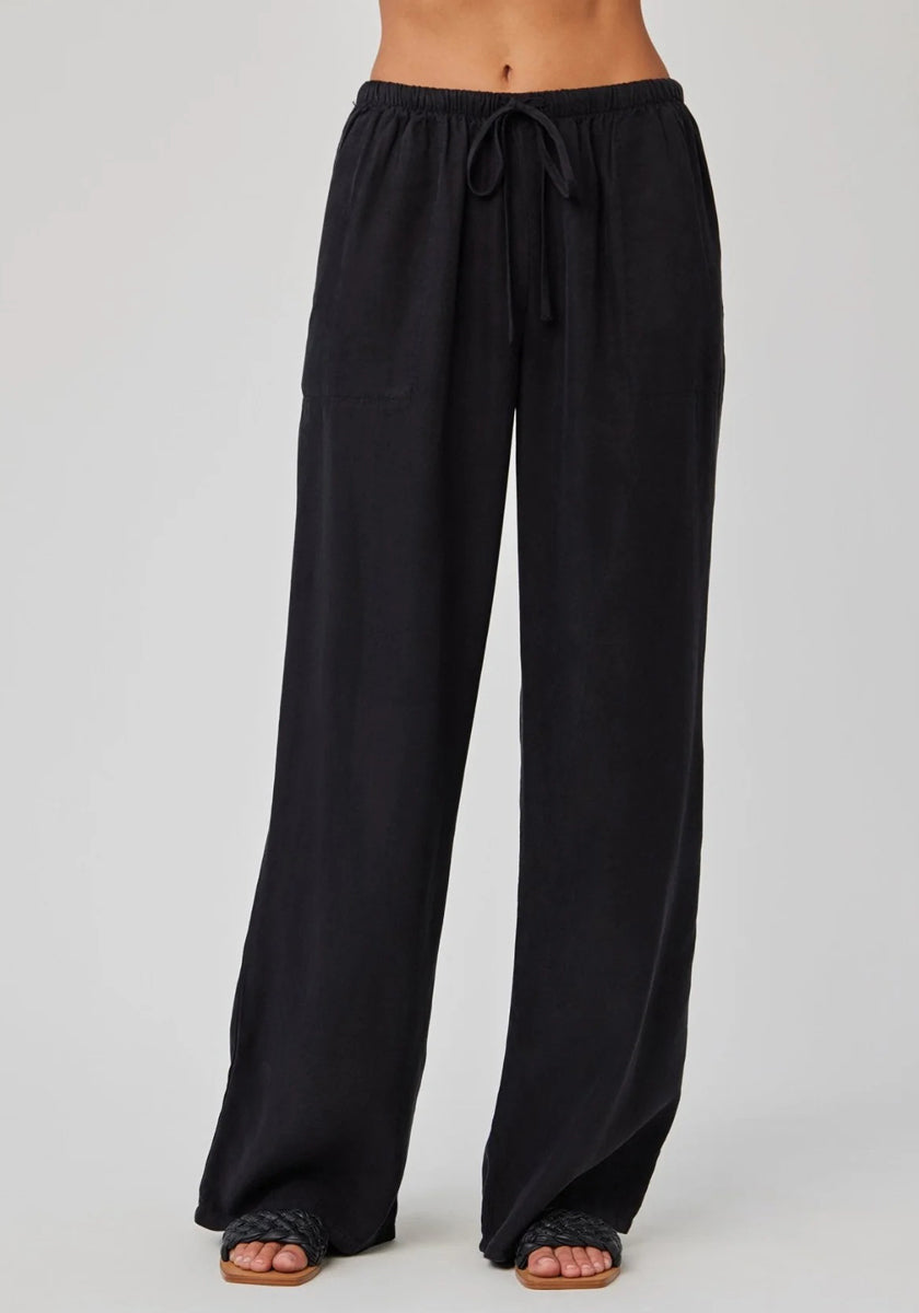 A model showcases Bella Dahl’s loose-fitting, high-waisted Lyocell pants with an elastic waistband, wide legs, and large side pockets. The cropped photo (midriff down) shows them barefoot against a light background.