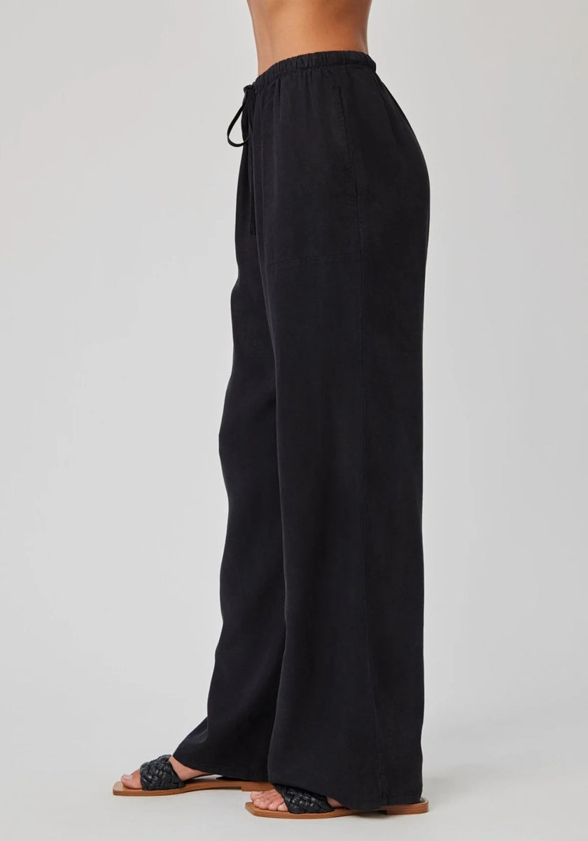 Eine Person trägt eine weite Lyocell-Hose mit elastischem Bund von Bella Dahl sowie schwarze, gewebte Slide-Sandalen. Die Seitenansicht vor hellgrauem Hintergrund betont den lässigen und bequemen Look des Outfits.
