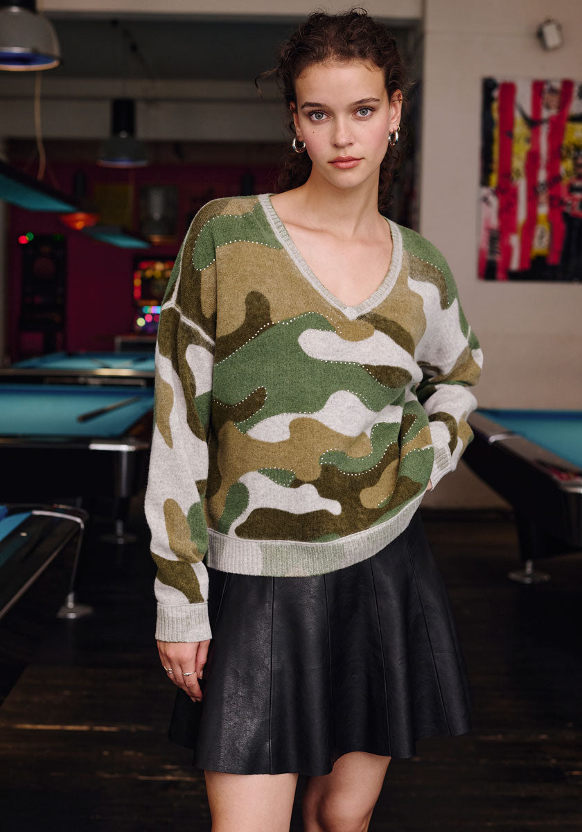 A young woman stands by pool tables, wearing a Frogbox by Princess loose camouflage V-neck sweater and a black faux leather miniskirt. Her curly dark hair is pulled back; she wears minimal jewelry and looks calm, with one hand in her skirt pocket.