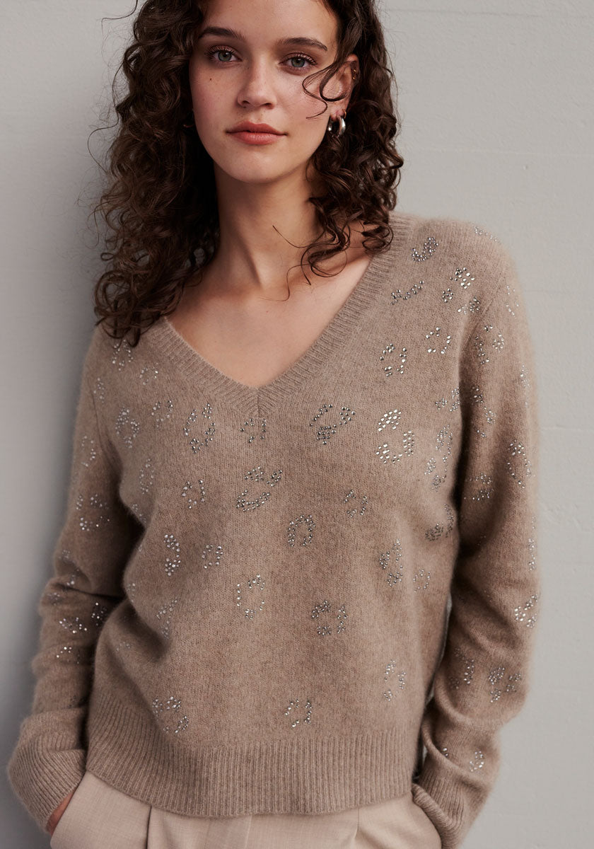 A young woman with wavy brown hair wears a beige Strickpullover mit V-Ausschnitt und funkelnder Strassverzierung from Princess Goes Hollywood. She stands against a light gray wall, hands in the pockets of her light-colored trousers.