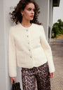A woman with curly brown hair stands outdoors near greenery, wearing a cream Fake Fur Jacke with gold buttons and brown leopard print pants from Princess Goes Hollywood. She holds a black handbag and gazes to the side by a white wall.