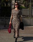 Eine Frau geht in einem wadenlangen Kleid mit Leopardenmuster, schwarzen klobigen Stiefeln und der Oversized Sonnenbrille von Okkia im Freien spazieren. Sie trägt eine rote, strukturierte Handtasche und eine schwarze Jacke; im Hintergrund sind Eisentore und Palmen zu sehen.