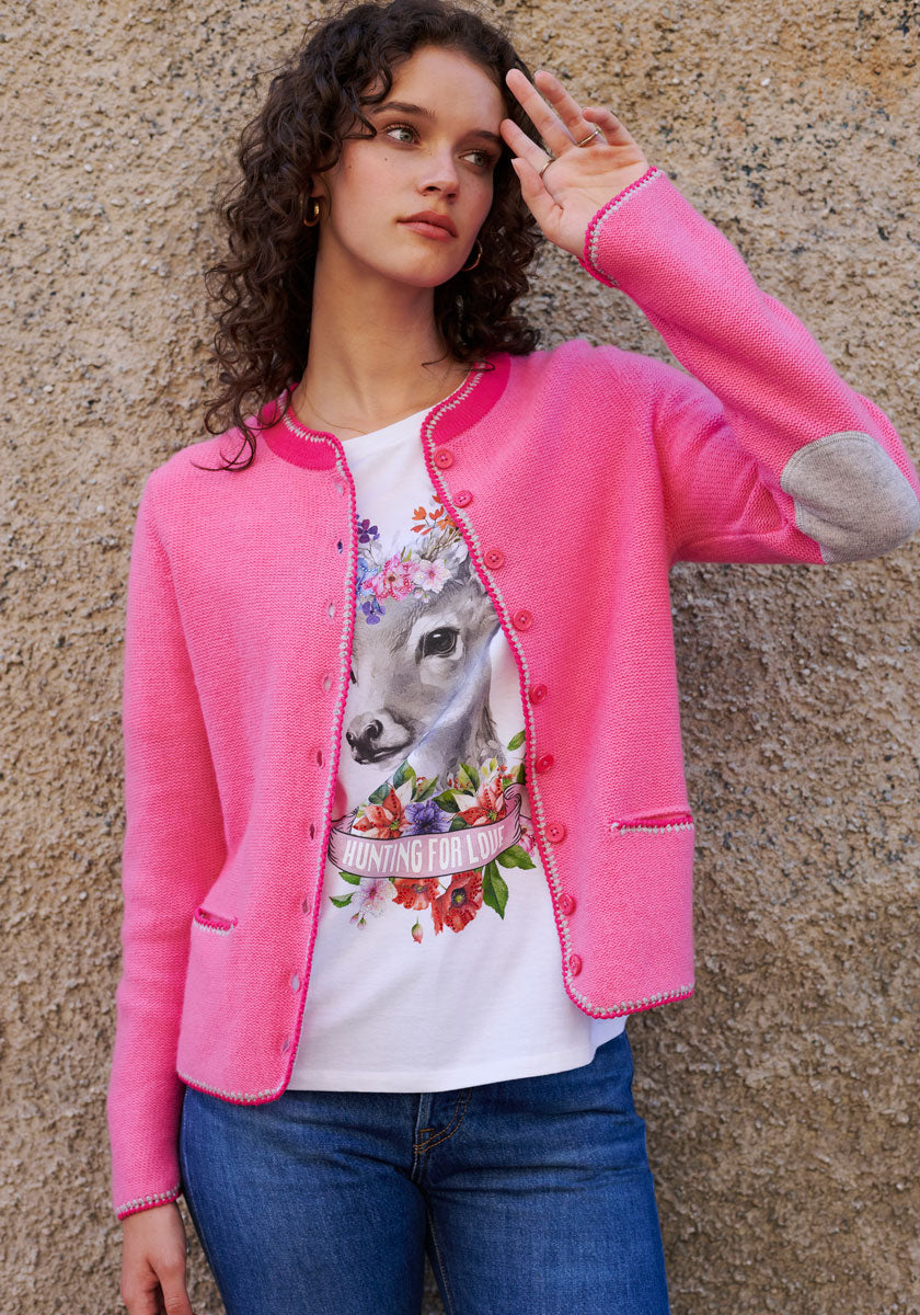 A young woman with curly hair stands by a textured beige wall, wearing a bright pink Princess Goes Hollywood Cardigan mit Strassverzierung and a white Baumwoll-T-Shirt with a Reh-Motiv, flower crown, and Hunting for Love text, gazing into the distance.