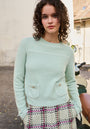 A young woman with curly brown hair wears a mint green Princess Goes Hollywood merino-cashmere blend pullover with a round neckline and a pink, black, and cream plaid skirt. She stands on a cobblestone street, leaning forward with a gentle expression.