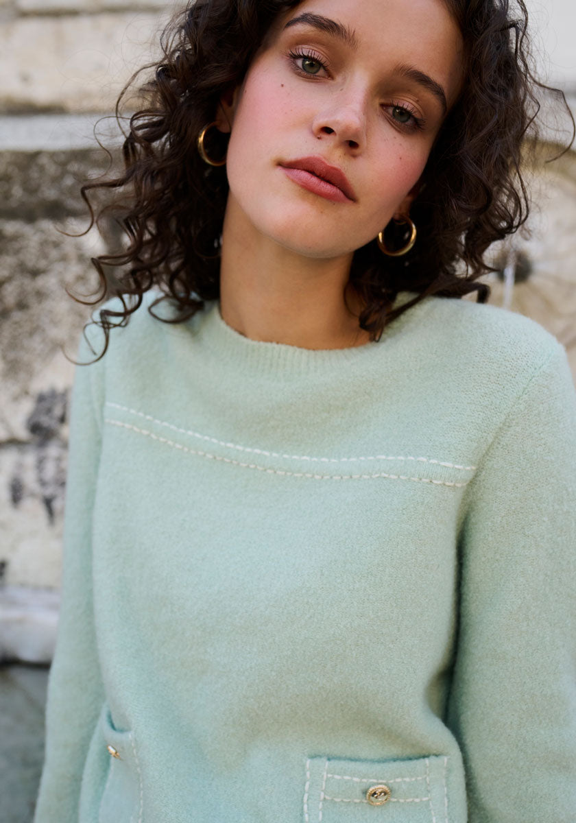 A young woman with curly brown hair and gold hoop earrings wears a mint green Princess Goes Hollywood Merino-Cashmere blend crewneck sweater with stitched details and pockets, standing outside against a stone wall and gazing calmly at the camera.