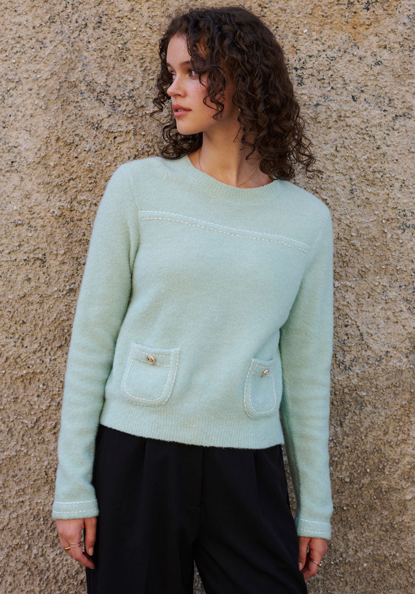 A woman with curly brown hair wears a mint-green Princess Goes Hollywood Strickpullover with Rundhalsausschnitt and two small front pockets, paired with black pants. She stands before a textured beige wall, gazing left relaxedly.