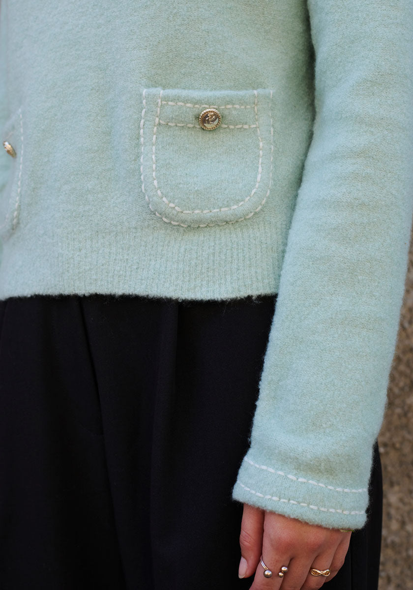 A person wears a light mint green Princess Goes Hollywood Strickpullover with Rundhalsausschnitt, patch pocket, and gold button detail, paired with high-waisted black pants. Their ring-adorned hand rests at their side before a blurred background.