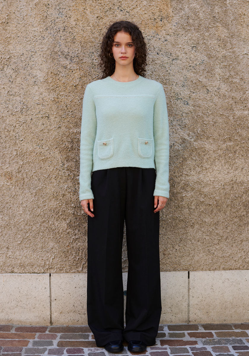 A woman with curly brown hair stands against a beige wall, wearing a Princess Goes Hollywood Merino-cashmere blend sweater with crew neck and two front pockets, paired with black wide-leg trousers. Her arms rest relaxed at her sides on stone tiles.