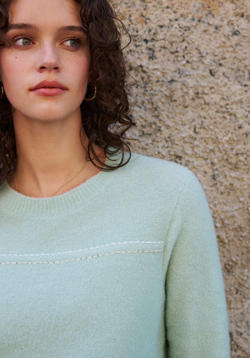 A young woman with curly brown hair and green eyes stands by a beige stone wall, wearing a mint green Princess Goes Hollywood Merino-Cashmere blend pullover with a crew neck and gold hoop earrings.