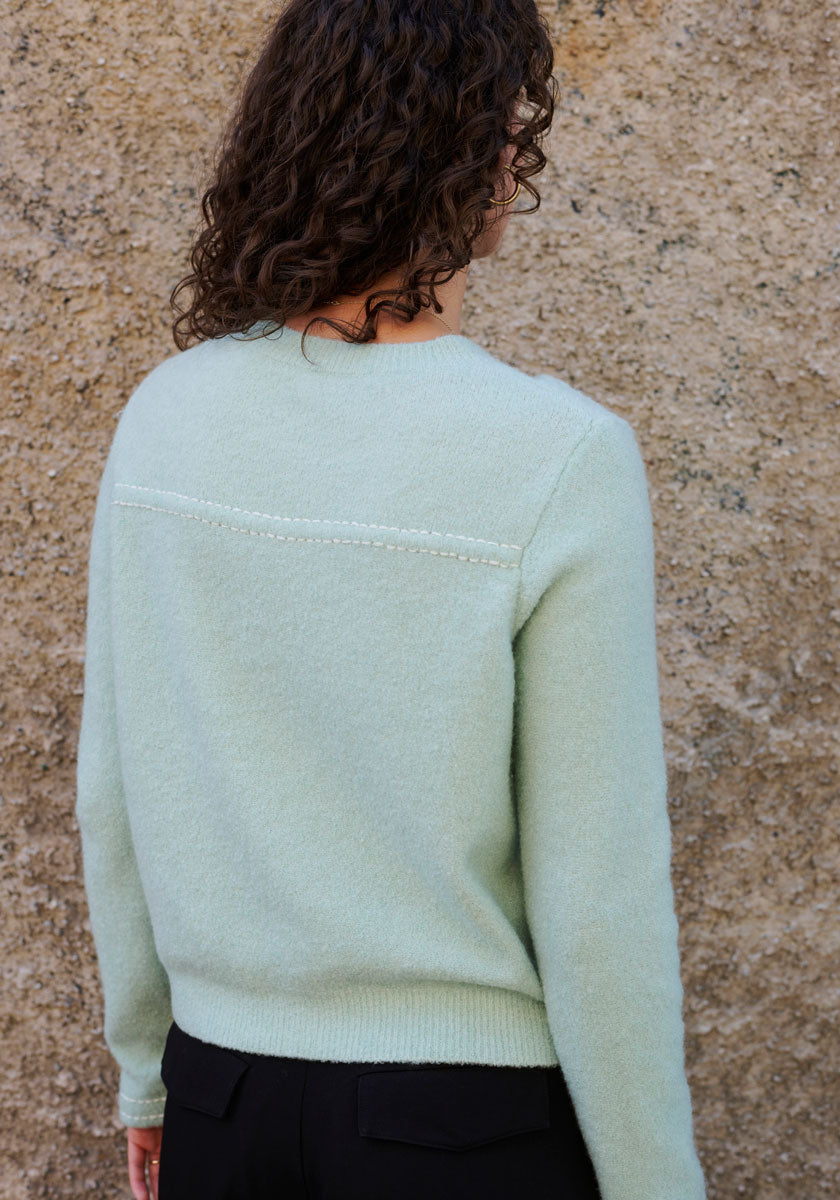 A person with curly brown hair, seen from behind, wears a light green Princess Goes Hollywood Merino-cashmere blend sweater with crew neck and black pants, standing before a rough tan wall.