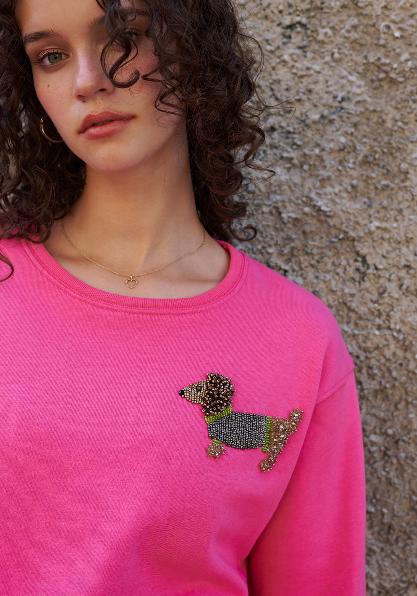 A young woman with curly brown hair wears a bright pink Princess Goes Hollywood sweatshirt featuring a Dackel motif with delicate pearl embroidery on the upper chest. She also wears a gold necklace and stands against a textured stone wall.