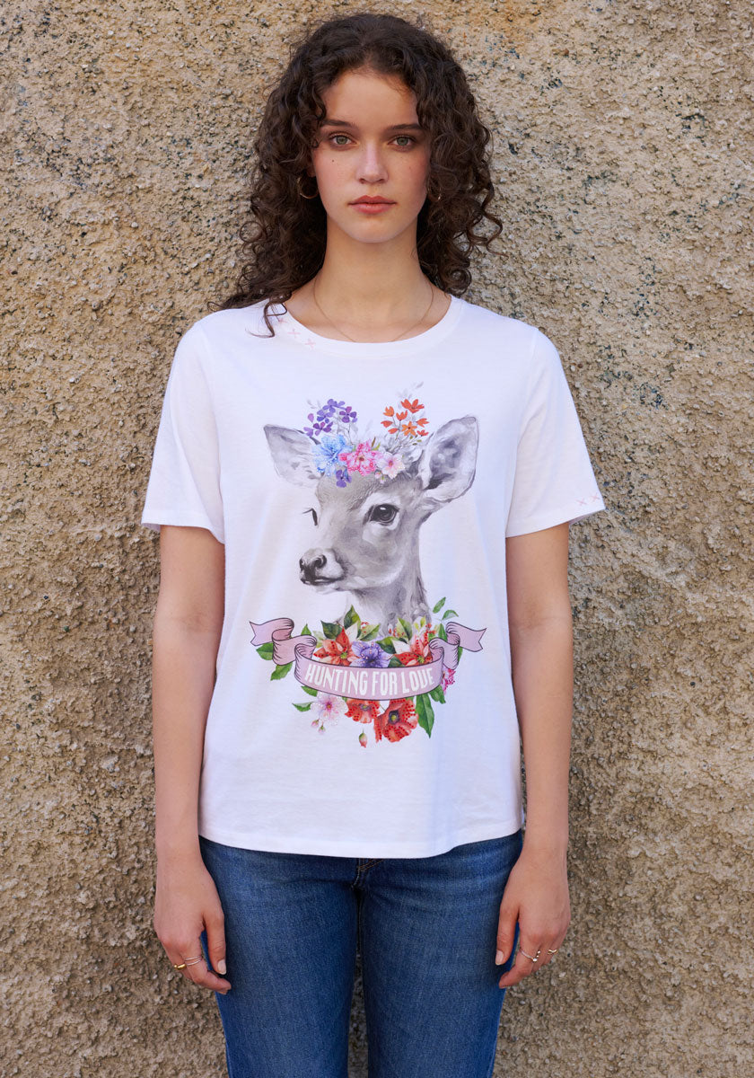 A young woman with curly brown hair stands by a beige stone wall, wearing Princess Goes Hollywood’s cotton T-shirt featuring a floral deer design and pink HUNTING FOR LOVE ribbon, paired with blue jeans. She gazes calmly into the camera.