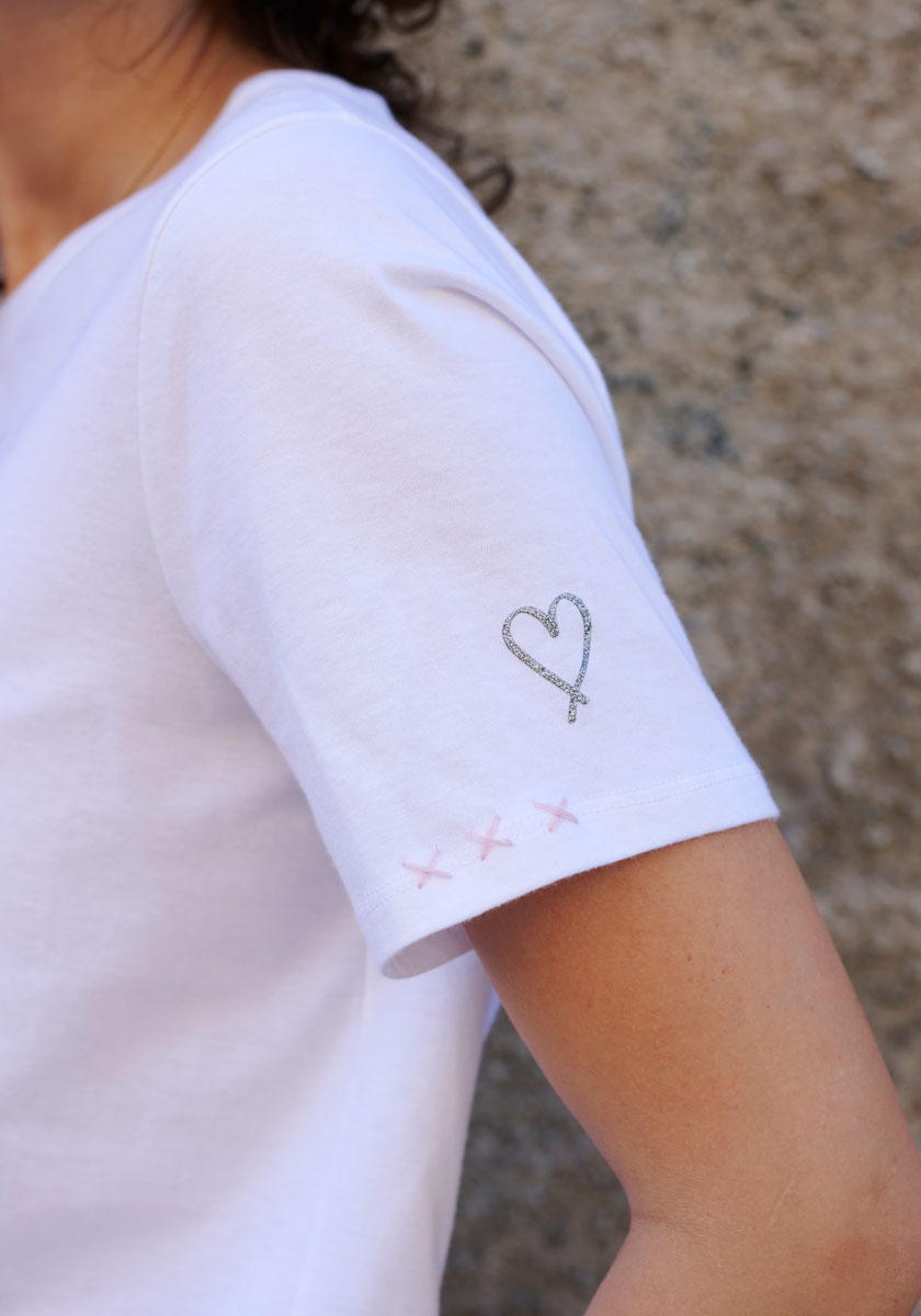 A person wearing a plain white Princess Goes Hollywood T-shirt is shown from the shoulder down. The shirt features a black outlined heart, three pink cross-stitches, and an “X” on the sleeve, set against a textured stone wall.