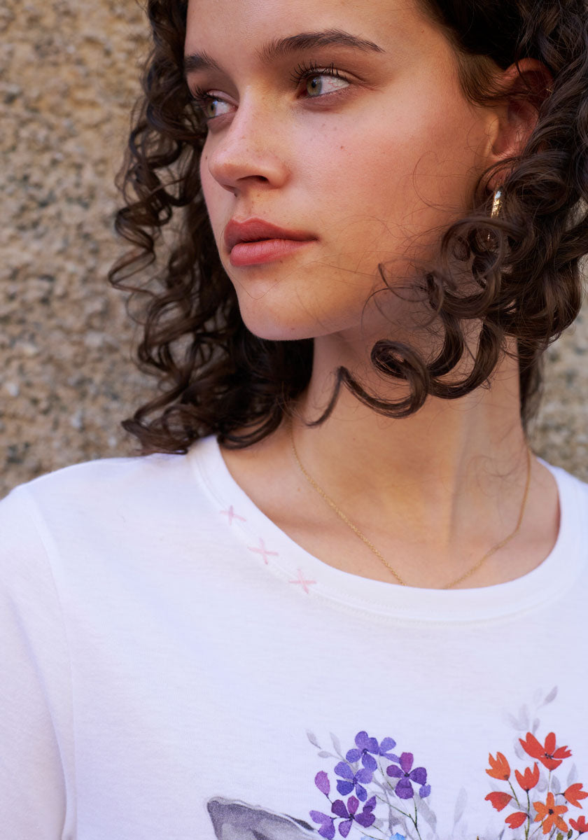 A young woman with curly brown hair and green eyes gazes to the side, wearing a white floral print Baumwolle shirt by Princess Goes Hollywood, a thin gold necklace, and small gold hoop earrings against a textured beige wall.