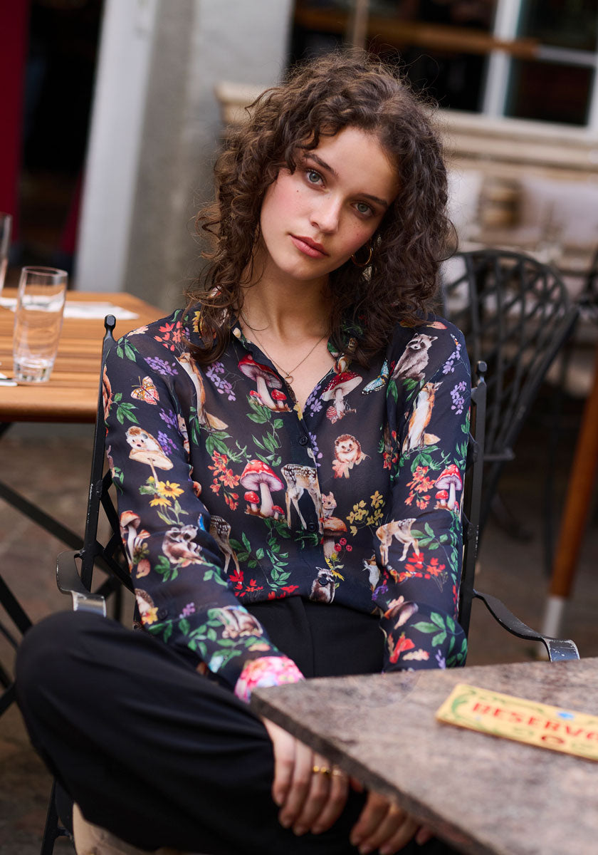 A young woman with curly brown hair sits outdoors, wearing a dark, transparent Princess Goes Hollywood Hemdbluse with colorful botanical and Waldtier-Prints, paired with black pants. She gazes softly at the camera, one hand resting casually on her lap.