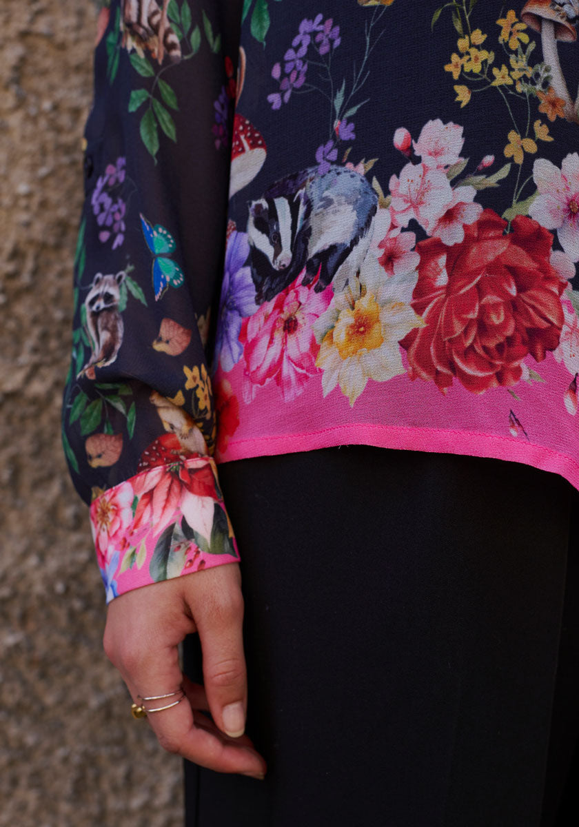 A person’s left hand with gold rings rests by their side, wearing Princess Goes Hollywood’s transparent long-sleeved Hemdbluse with a colorful badger and floral Waldtier-Print, pink hem, black pants, and a stone background.