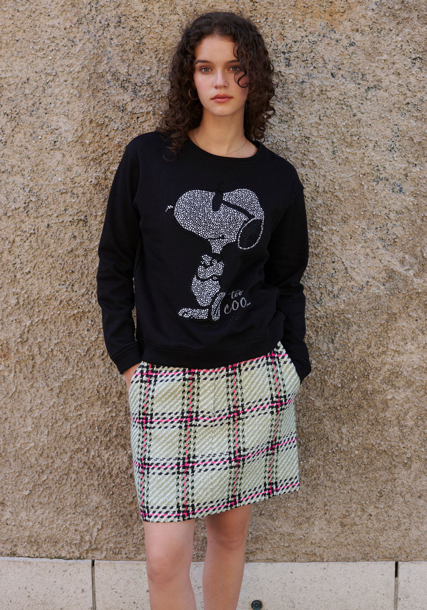 A young woman with curly brown hair stands against a textured beige wall, wearing a Princess Goes Hollywood black Sweatshirt Damen with playful Snoopy-Print und Strassverzierung and a high-waisted, pleated plaid skirt in light green, white, and red. Her hands are in her pockets.