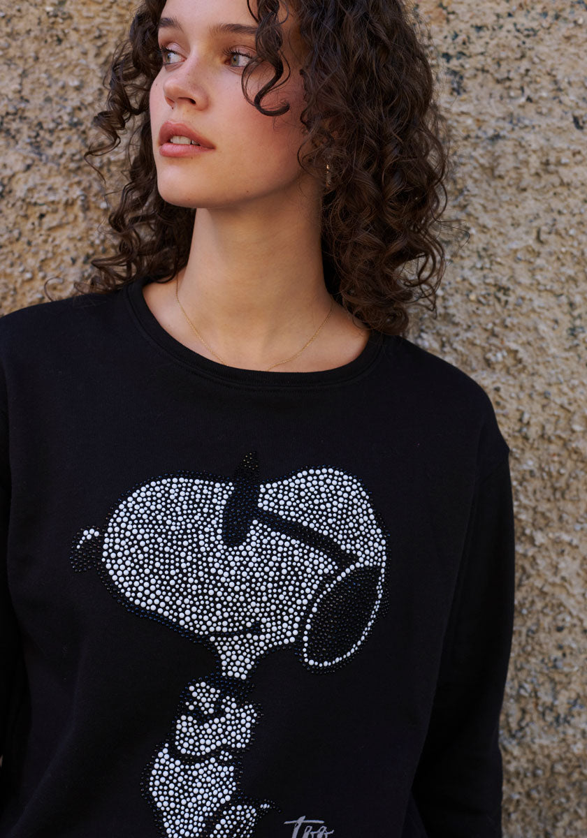 A young woman with curly brown hair stands by a stone wall, wearing a black Princess Goes Hollywood Snoopy Sweatshirt aus Baumwolle with a white strassverzierung graphic. Soft natural light highlights her upper body as she gazes to the side.