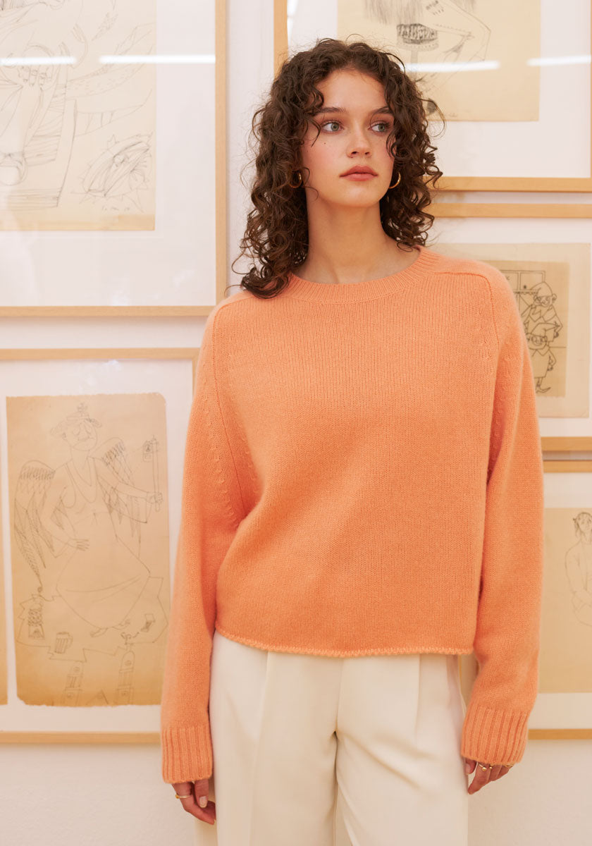 A woman with curly brown hair wears a loose, peach-colored Princess Goes Hollywood Kaschmir Pullover mit Rundhalsausschnitt and cream pants. She stands before framed sketches, gazing slightly right, her expression neutral and arms relaxed at her sides.
