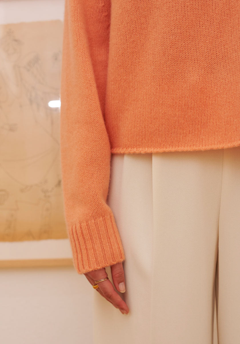 A person wears a peach Kaschmir Pullover with Rundhalsausschnitt and long sleeves by Princess Goes Hollywood, paired with cream pants. Only their torso and part of one arm with rings are visible; a framed sketch hangs behind them.