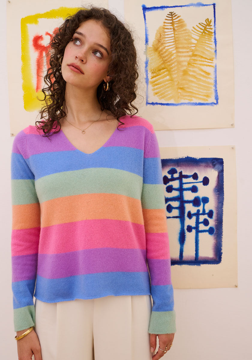 A young woman with curly brown hair wears a Princess Goes Hollywood gestreifter Pullover in pastel rainbow hues and cream pants, gazing upward pensively. Behind her, abstract art features a red figure, golden fern leaves, and blue patterns on off-white paper.