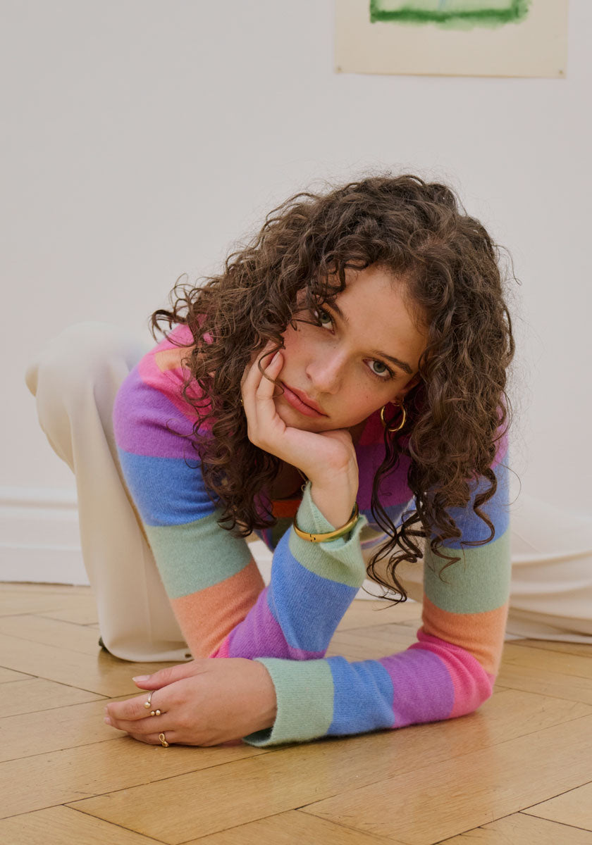 A young woman with curly brown hair crouches indoors on a wooden floor, thoughtfully gazing at the camera. She wears a pastel striped pullover and cream pants from Princess Goes Hollywood, set against pale walls and minimal art.