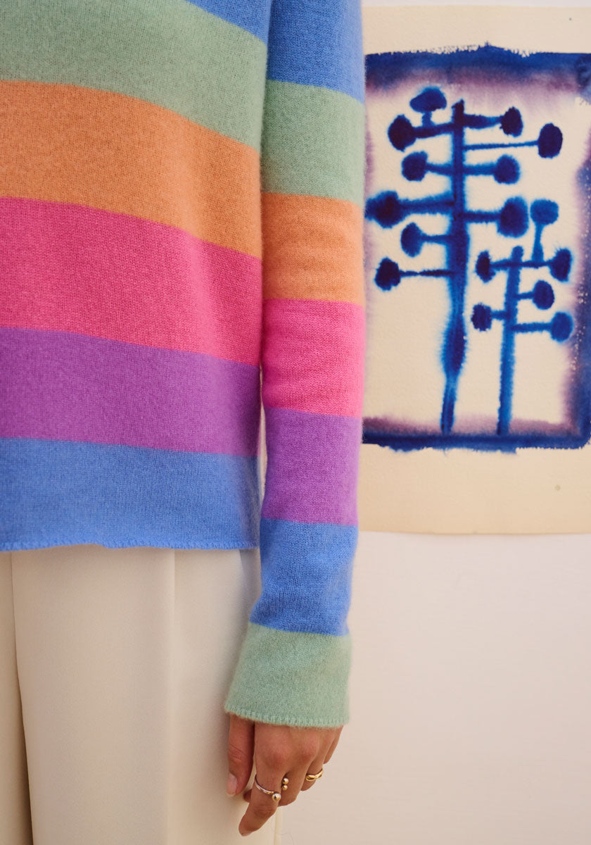 A close-up of a person in a Princess Goes Hollywood striped pullover (blue, green, pink, purple, orange) and beige pants. Their rings are visible. Abstract blue tree-like artwork with round dots appears in the background.