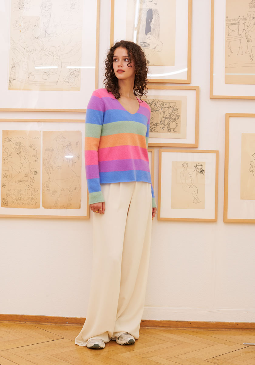 A woman with curly hair wears a Princess Goes Hollywood gestreifter Pullover in pastel rainbow tones, wide-leg cream pants, and white sneakers. She stands indoors on a wooden floor by framed sketches, gazing slightly to the side.