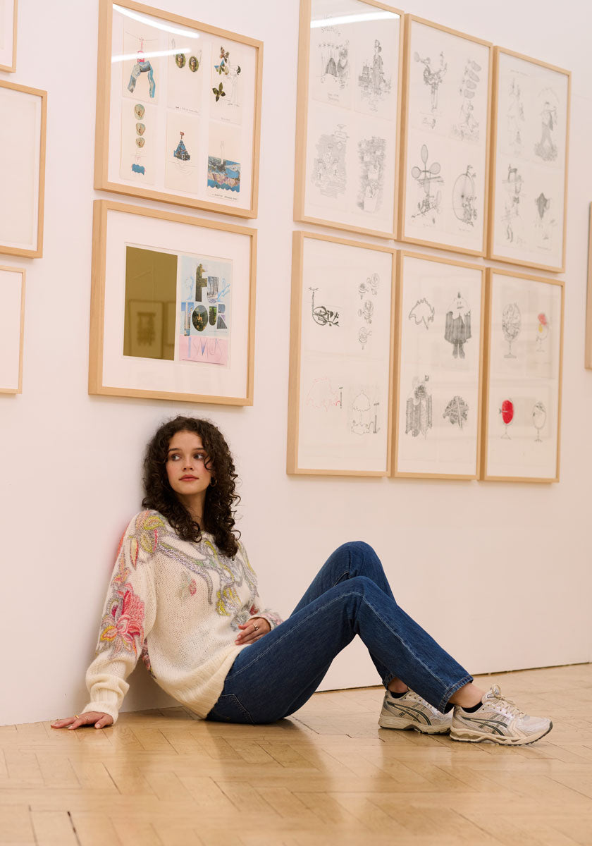 A woman with curly hair, wearing a Princess Goes Hollywood Pullover Wolle-Alpaka-Mix with colorful floral designs, blue jeans, and sneakers, sits on a wooden floor against a white wall lined with framed art sketches.