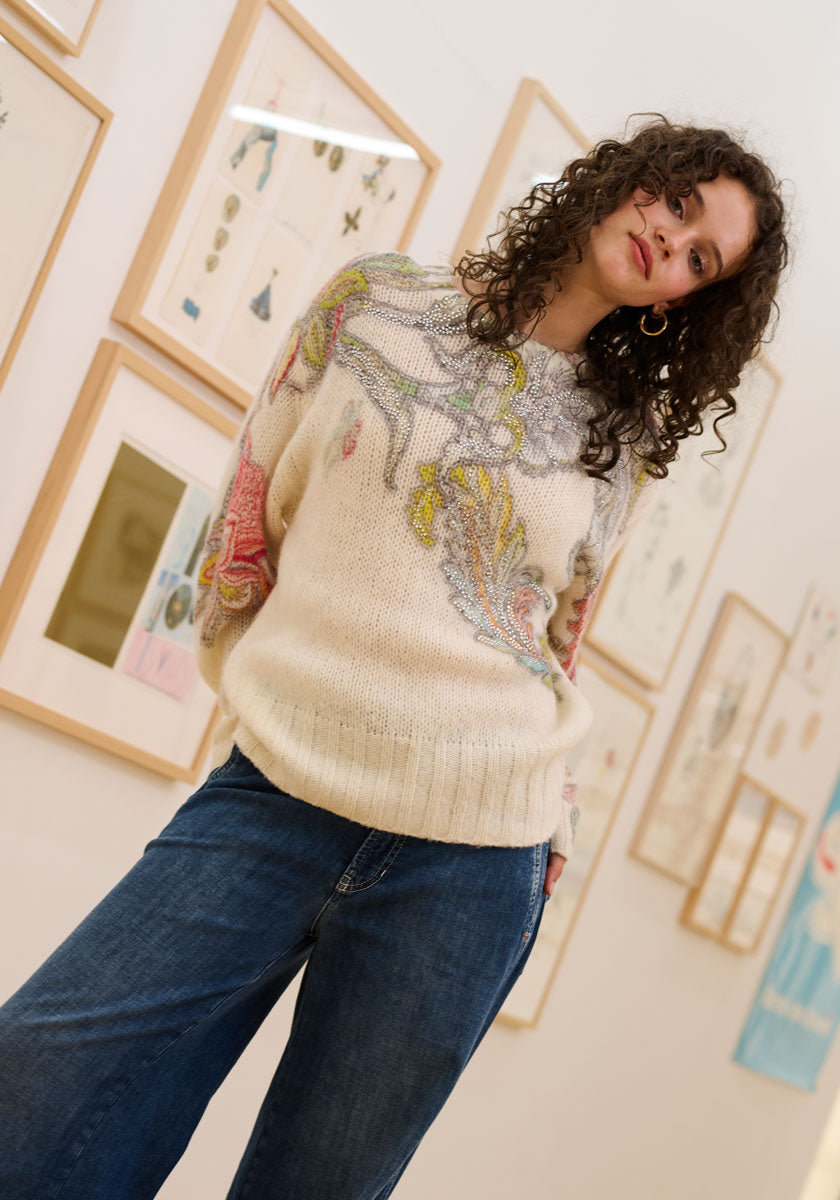 A woman with curly dark hair wears a cream Princess Goes Hollywood Pullover Wolle-Alpaka-Mix with colorful embroidery and wide-leg blue jeans, standing before framed art and botanical prints with a gentle, relaxed expression.