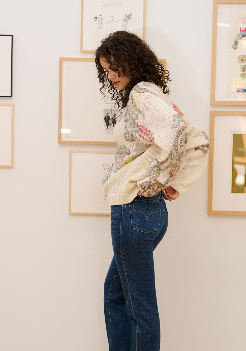 A woman with curly brown hair stands in profile, looking down, wearing a cream Princess Goes Hollywood Pullover Wolle-Alpaka-Mix with paisley design and blue jeans. She is in front of a white wall decorated with framed art in light wooden frames.