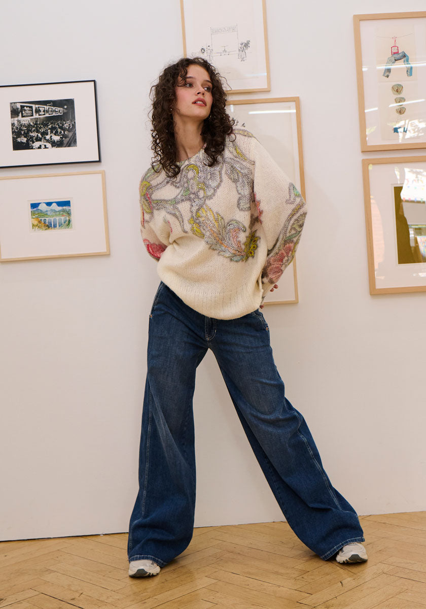 A woman with curly brown hair wears a loose Princess Goes Hollywood Wolle-Alpaka-Mix Pullover with a colorful floral pattern, wide-leg blue jeans, and sneakers. She stands indoors on a wooden floor; framed art decorates the wall behind her.