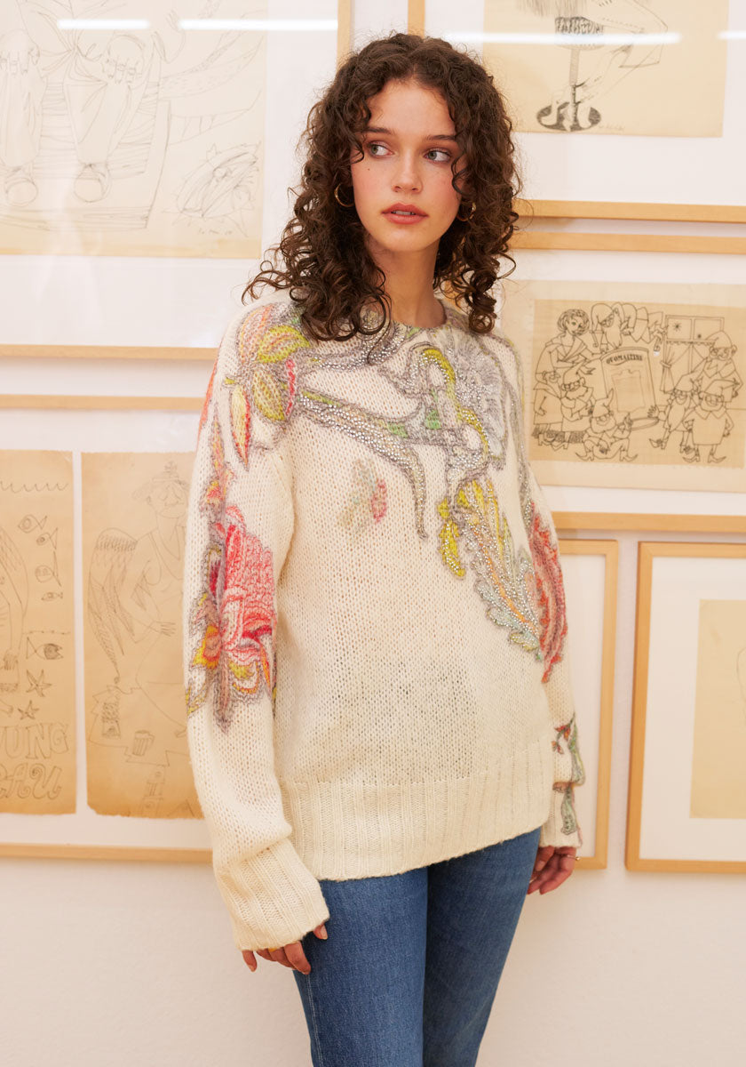 A young woman with curly brown hair wears a cream Princess Goes Hollywood Wolle-Alpaka-Mix Pullover, featuring large colorful floral embroidery on the sleeves and chest, standing indoors before a wall of framed sketches and looking to her left.
