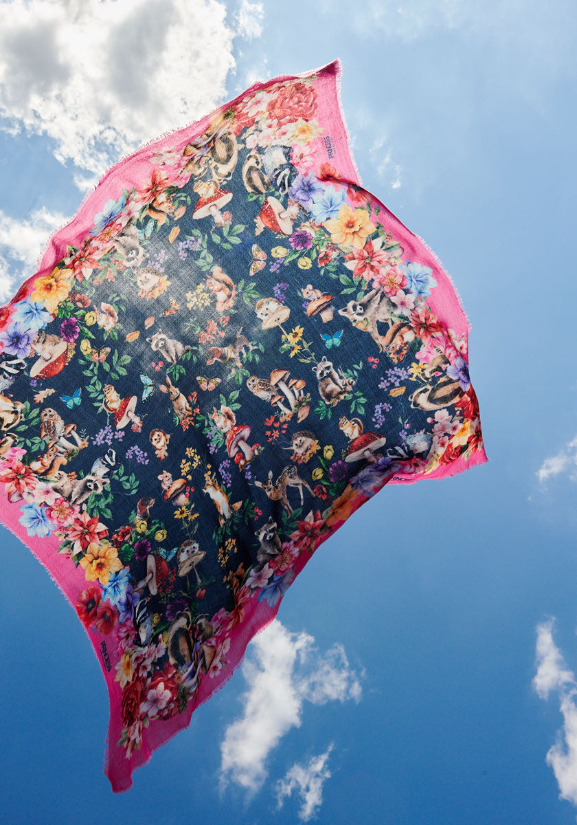 A colorful quadratischer Schal by Princess Goes Hollywood, featuring a vibrant floral and Waldtier-Print with bright pink borders, floats mid-air against a blue sky and fluffy clouds, evoking movement and lightness.
