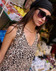 A woman in a Princess Goes Hollywood floral leopard-print silk maxi dress, large black sunglasses, and a black headscarf stands outside a market stall with fruits in the background, accessorized with a gold chain necklace.