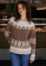 A woman with curly brown hair and gold hoop earrings stands indoors by a coffee shop counter, wearing a cozy Princess Goes Hollywood Jacquardpullover with Raglanärmel in a Wolle-Alpaka-Mix, paired with blue jeans.